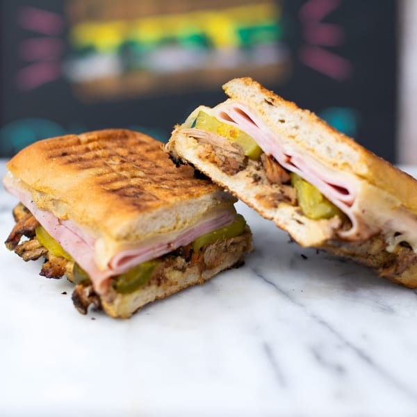 Two halves of a grilled Cuban sandwich with ham, pork, cheese, and pickles on a marble surface.