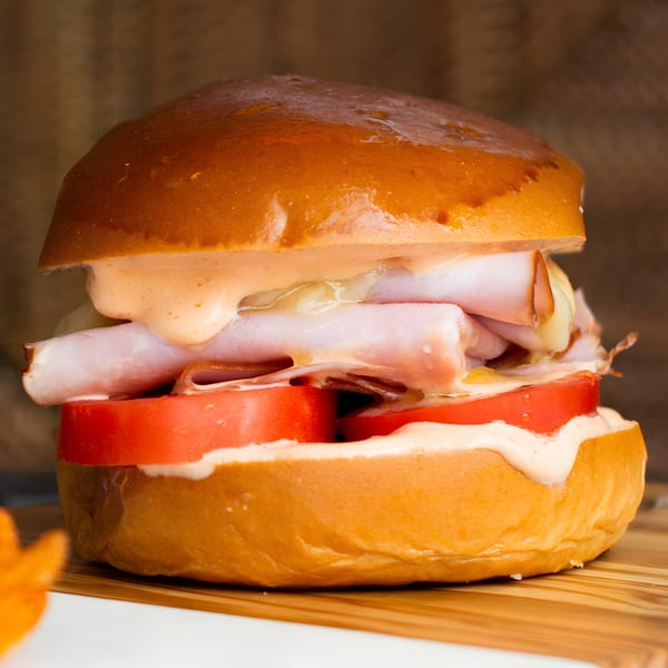 A close-up of a Sandwich Shop sandwich with ham, cheese, tomatoes, and sauce on a brioche bun.