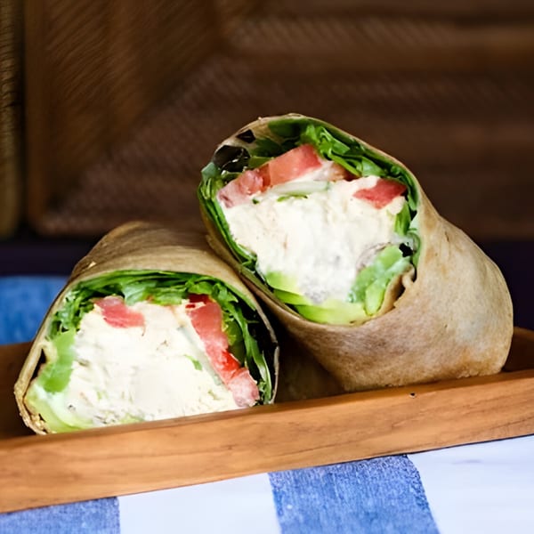 Two halves of a chicken salad wrap with lettuce, tomato, and avocado on a wooden tray.
